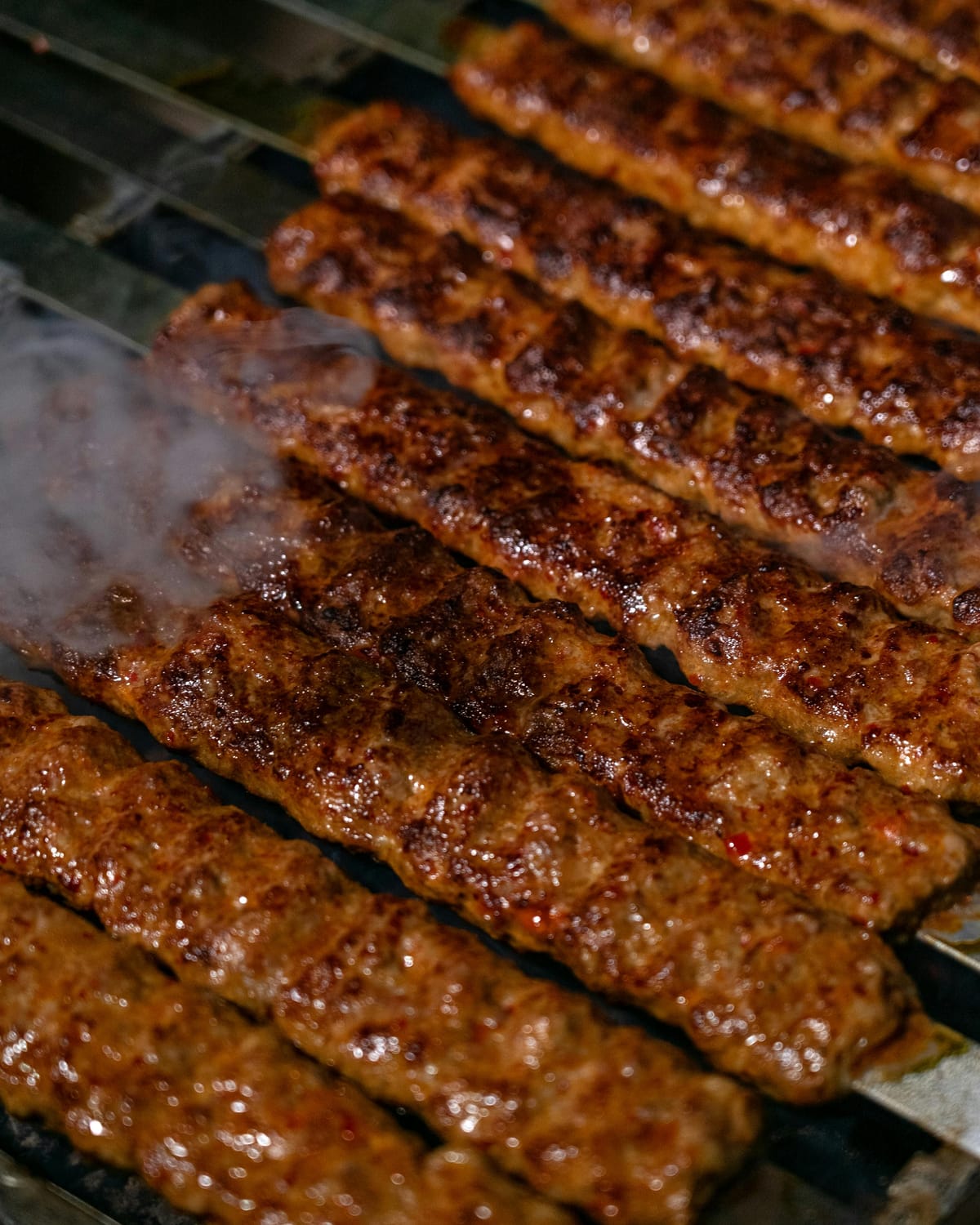 Close-up of Koobideh kababs grilling on flat skewers, perfectly intact, with steam rising